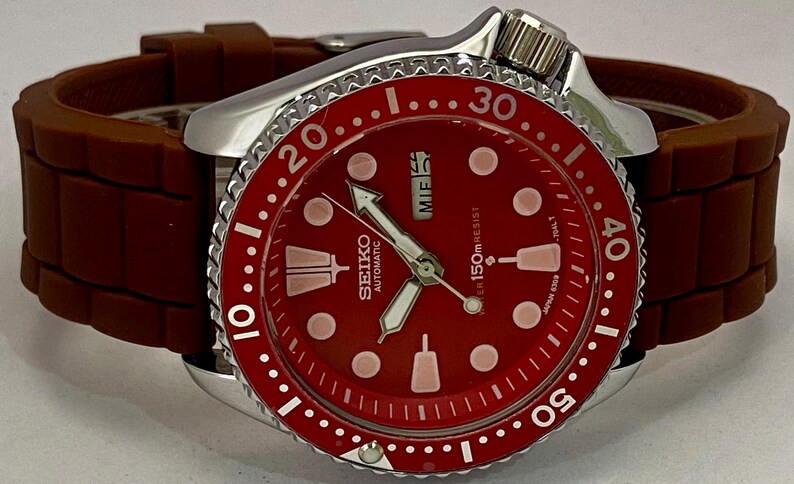 K&ouml;nnte beinhalten: Seiko Automatik-Armbanduhr mit rotem Zifferblatt und braunem Kautschukarmband. Die Uhr verf&uuml;gt &uuml;ber eine silberne L&uuml;nette mit wei&szlig;en Markierungen und eine Datumsanzeige. Das Zifferblatt hat wei&szlig;e Zeiger und Markierungen.