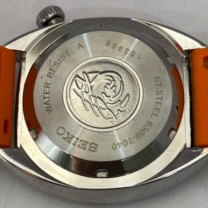 K&ouml;nnte beinhalten: Nahaufnahme einer silberfarbenen Seiko-Uhr mit orangefarbenem Armband. Auf der R&uuml;ckseite der Uhr sind das Seiko-Logo, die Worte "WATER RESIST" und "ST. STEEL" sowie die Zahlen "152956" und "6309-7040" zu sehen.