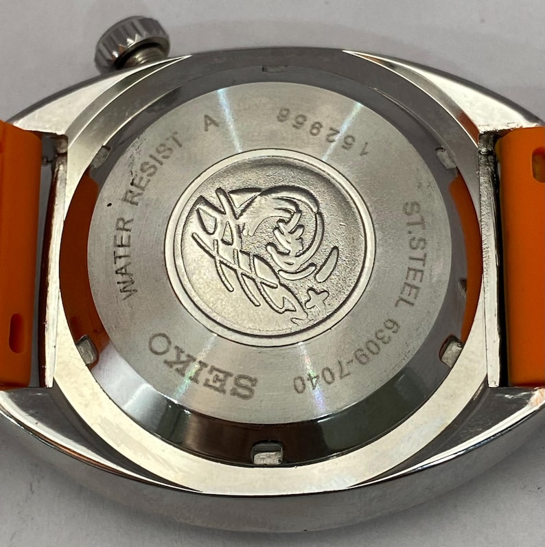 K&ouml;nnte beinhalten: Nahaufnahme einer silberfarbenen Seiko-Uhr mit orangefarbenem Armband. Auf der R&uuml;ckseite der Uhr sind das Seiko-Logo, die Worte "WATER RESIST" und "ST. STEEL" sowie die Zahlen "152956" und "6309-7040" zu sehen.