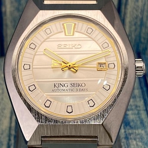 May include: A silver-toned King Seiko automatic wristwatch with a white dial and gold-colored hands and markers. The watch face displays the Seiko logo and the text "KING SEIKO AUTOMATIC 3 DAYS". The watch has a rectangular case and a silver-toned band.