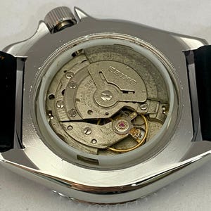 May include: Close-up of a silver-toned Seiko watch with a black leather strap. The watch's intricate internal mechanism is visible through the clear back, showcasing gears, springs, and the Seiko brand name.