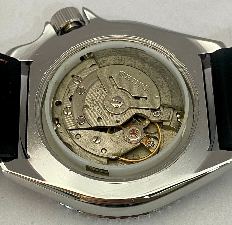 May include: Close-up of a silver-toned Seiko watch with a black leather strap. The watch's intricate internal mechanism is visible through the clear back, showcasing gears, springs, and the Seiko brand name.