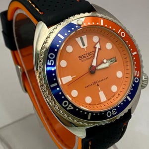 K&ouml;nnte beinhalten: Eine Seiko Automatik-Uhr aus Edelstahl mit orangefarbenem Zifferblatt, blau-oranger L&uuml;nette und schwarzem Kautschukarmband. Das Zifferblatt zeigt den Markennamen und "Water 150m Resist". Das Armband hat orangefarbene Akzente.
