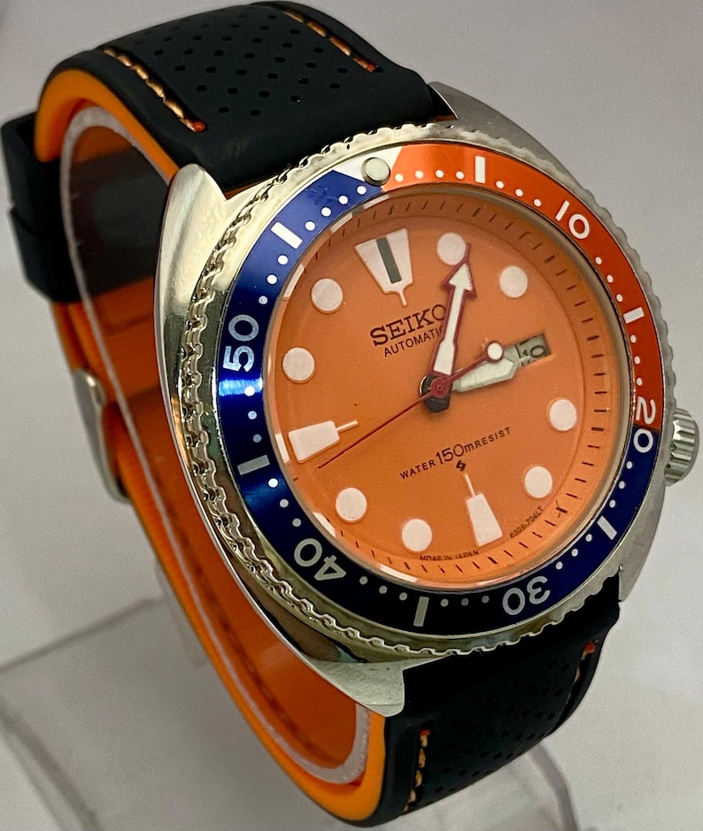 K&ouml;nnte beinhalten: Eine Seiko Automatik-Uhr aus Edelstahl mit orangefarbenem Zifferblatt, blau-oranger L&uuml;nette und schwarzem Kautschukarmband. Das Zifferblatt zeigt den Markennamen und "Water 150m Resist". Das Armband hat orangefarbene Akzente.