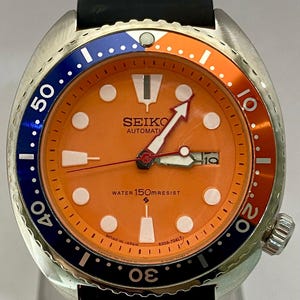 K&ouml;nnte beinhalten: Seiko Automatik-Uhr mit orangefarbenem Zifferblatt, blau-oranger L&uuml;nette und schwarzem Armband. Das Zifferblatt zeigt das Datum und den Text "WATER 150mRESIST".