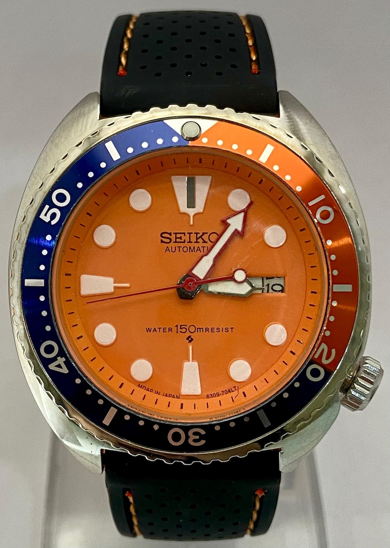 K&ouml;nnte beinhalten: Seiko Automatik-Uhr mit orangefarbenem Zifferblatt, blau-oranger L&uuml;nette und schwarzem Armband. Das Zifferblatt zeigt das Datum und den Text "WATER 150mRESIST".