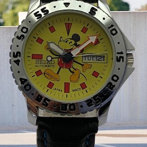 May include: A Seiko automatic wristwatch with a yellow face featuring a Mickey Mouse graphic. The watch has a silver bezel and a black leather band with blue stitching. The watch face displays the day and date.