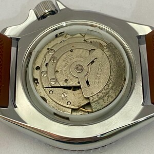 K&ouml;nnte beinhalten: Nahaufnahme einer Vintage Seiko-Uhr mit silberfarbenem Geh&auml;use und braunem Lederarmband. Der Uhrenr&uuml;cken ist ge&ouml;ffnet und zeigt das komplizierte mechanische Uhrwerk, einschlie&szlig;lich Zahnr&auml;dern, Juwelen und dem Markennamen "SEIKO". Das Zifferblatt ist sichtbar.