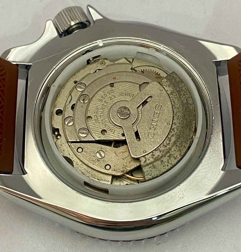 K&ouml;nnte beinhalten: Nahaufnahme einer Vintage Seiko-Uhr mit silberfarbenem Geh&auml;use und braunem Lederarmband. Der Uhrenr&uuml;cken ist ge&ouml;ffnet und zeigt das komplizierte mechanische Uhrwerk, einschlie&szlig;lich Zahnr&auml;dern, Juwelen und dem Markennamen "SEIKO". Das Zifferblatt ist sichtbar.