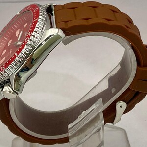 K&ouml;nnte beinhalten: Eine Edelstahl-Armbanduhr mit rotem Zifferblatt und braunem Silikonarmband. Die Uhr hat eine rote L&uuml;nette mit wei&szlig;en Markierungen und ein silbernes Geh&auml;use. Das Armband ist strukturiert und sicher befestigt.