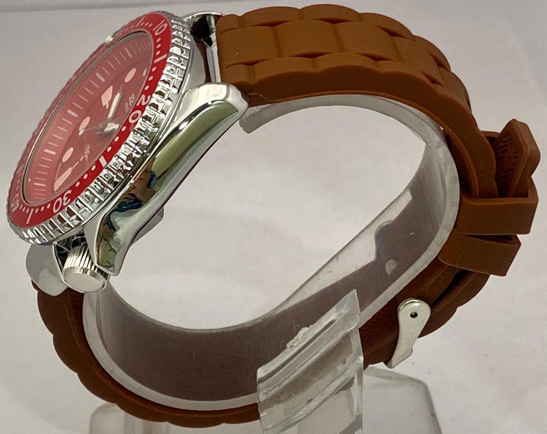 K&ouml;nnte beinhalten: Eine Edelstahl-Armbanduhr mit rotem Zifferblatt und braunem Silikonarmband. Die Uhr hat eine rote L&uuml;nette mit wei&szlig;en Markierungen und ein silbernes Geh&auml;use. Das Armband ist strukturiert und sicher befestigt.