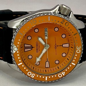 May include: A Seiko automatic watch with an orange dial and a black leather strap. The watch features a silver bezel with white markings and a date display at the 3 o'clock position. The watch is water resistant to 150m.