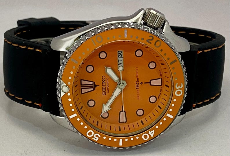 May include: A Seiko automatic watch with an orange dial and a black leather strap. The watch features a silver bezel with white markings and a date display at the 3 o'clock position. The watch is water resistant to 150m.
