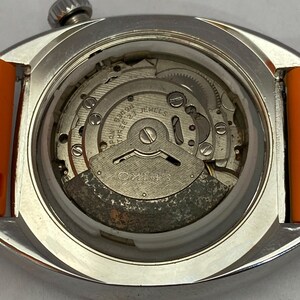 K&ouml;nnte beinhalten: Nahaufnahme einer Vintage Seiko-Uhr mit orangefarbenem Armband. Der Uhrenr&uuml;cken ist ge&ouml;ffnet und zeigt das komplizierte mechanische Uhrwerk mit Zahnr&auml;dern, Schrauben und dem Seiko-Logo. Die Uhr hat ein silberfarbenes Geh&auml;use.