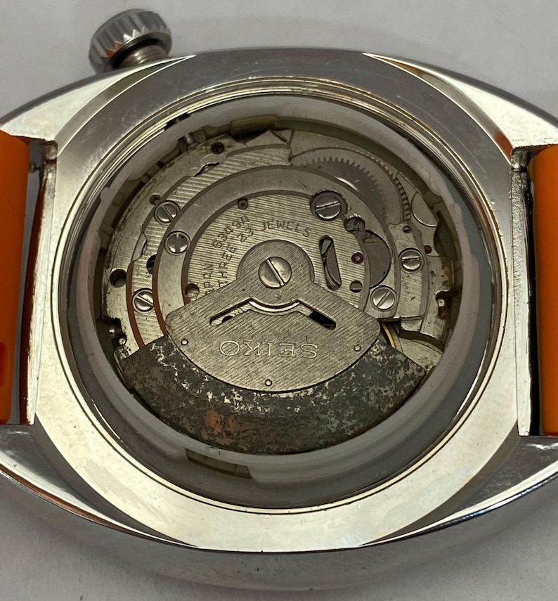 K&ouml;nnte beinhalten: Nahaufnahme einer Vintage Seiko-Uhr mit orangefarbenem Armband. Der Uhrenr&uuml;cken ist ge&ouml;ffnet und zeigt das komplizierte mechanische Uhrwerk mit Zahnr&auml;dern, Schrauben und dem Seiko-Logo. Die Uhr hat ein silberfarbenes Geh&auml;use.