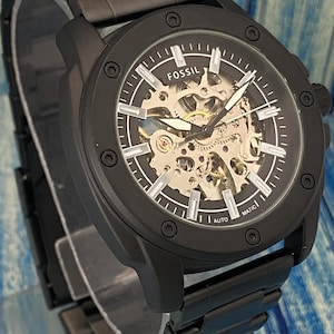 May include: Black Fossil automatic watch with a skeleton dial. The watch features a black metal band and case. The watch face displays the Fossil logo and the words "AUTO" and "MATIC". The watch has silver hands and markers.