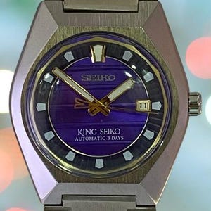 May include: A vintage Seiko King Seiko automatic wristwatch. The watch features a purple dial with gold hands and markers, a date window, and the text "KING SEIKO AUTOMATIC 3 DAYS". The watch has a silver-tone metal case and bracelet.