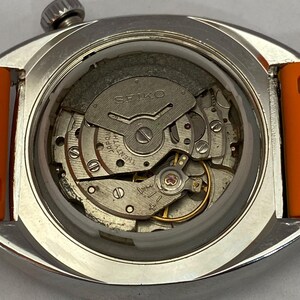 K&ouml;nnte beinhalten: Nahaufnahme einer Vintage Seiko-Uhr ohne R&uuml;ckseite, die das komplizierte mechanische Uhrwerk zeigt. Die Uhr hat ein silbernes Geh&auml;use und ein orangefarbenes Armband. Die inneren Komponenten, einschlie&szlig;lich Zahnr&auml;der und das Seiko-Logo, sind sichtbar.