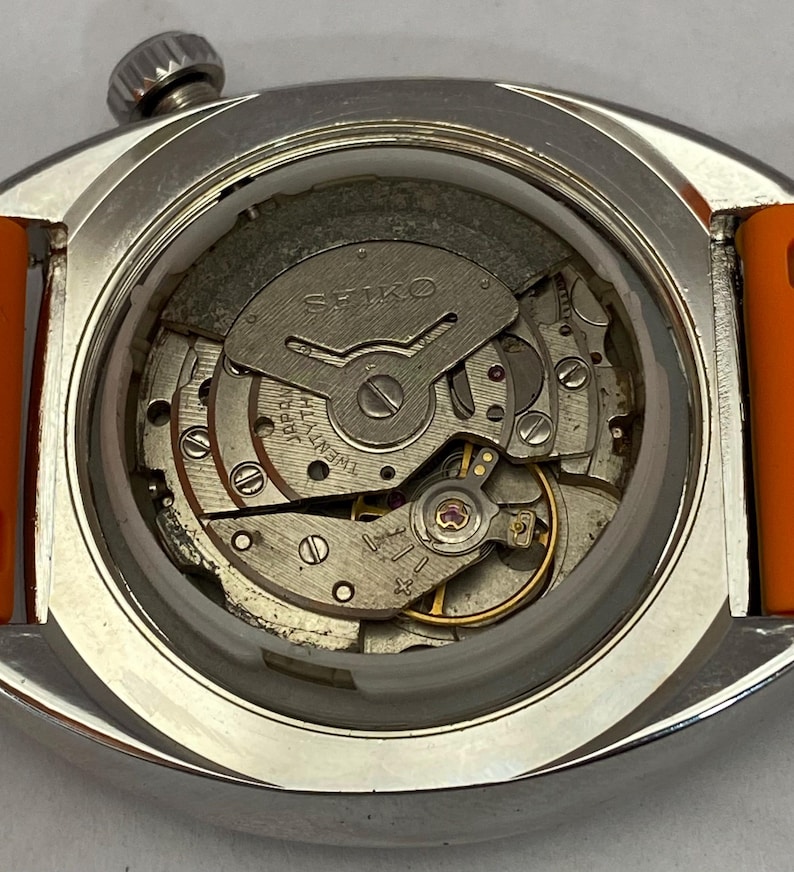 K&ouml;nnte beinhalten: Nahaufnahme einer Vintage Seiko-Uhr ohne R&uuml;ckseite, die das komplizierte mechanische Uhrwerk zeigt. Die Uhr hat ein silbernes Geh&auml;use und ein orangefarbenes Armband. Die inneren Komponenten, einschlie&szlig;lich Zahnr&auml;der und das Seiko-Logo, sind sichtbar.