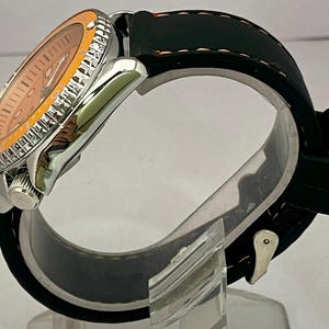 May include: A silver wristwatch with an orange dial and bezel. The watch has a black rubber strap. The watch is displayed on a clear stand. The watch face has silver hands and markers.