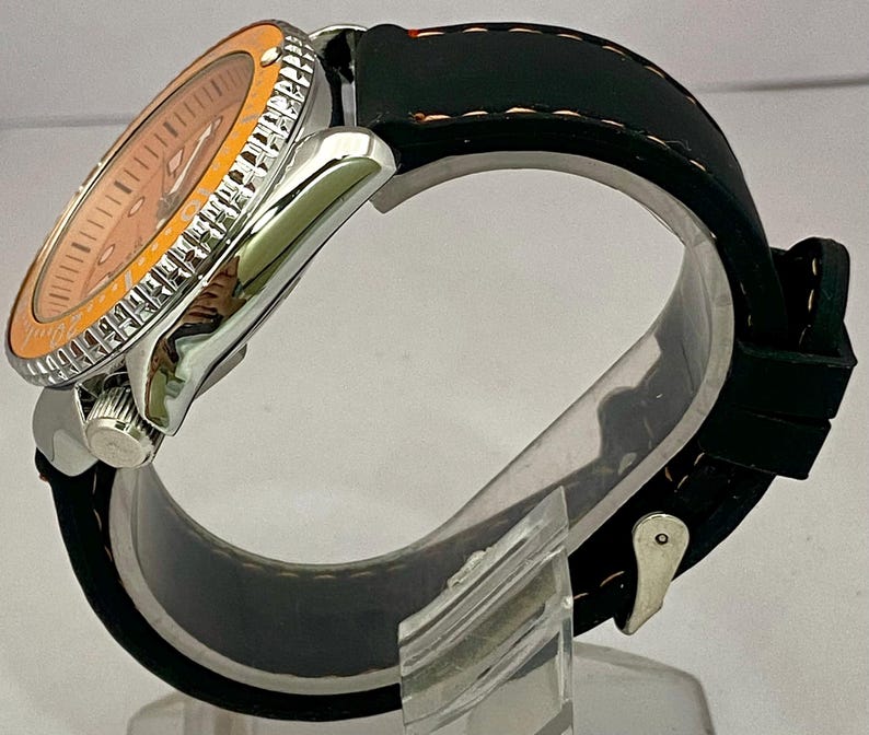 May include: A silver wristwatch with an orange dial and bezel. The watch has a black rubber strap. The watch is displayed on a clear stand. The watch face has silver hands and markers.