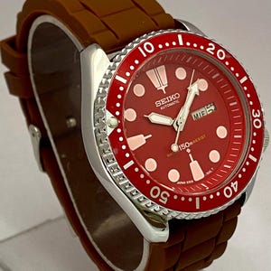 K&ouml;nnte beinhalten: Eine Seiko Automatik-Uhr aus Edelstahl mit rotem Zifferblatt und L&uuml;nette. Die Uhr hat ein braunes Kautschukarmband und eine Datumsanzeige. Das Zifferblatt hat wei&szlig;e Markierungen und Zeiger, mit dem Seiko-Logo.