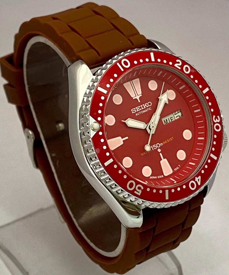 K&ouml;nnte beinhalten: Eine Seiko Automatik-Uhr aus Edelstahl mit rotem Zifferblatt und L&uuml;nette. Die Uhr hat ein braunes Kautschukarmband und eine Datumsanzeige. Das Zifferblatt hat wei&szlig;e Markierungen und Zeiger, mit dem Seiko-Logo.