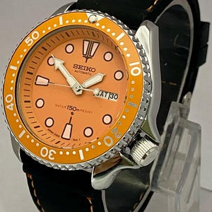 May include: A stainless steel Seiko automatic watch with an orange dial and bezel. The watch features a black leather strap with orange stitching. The dial displays the day and date. The watch is water resistant to 150m.