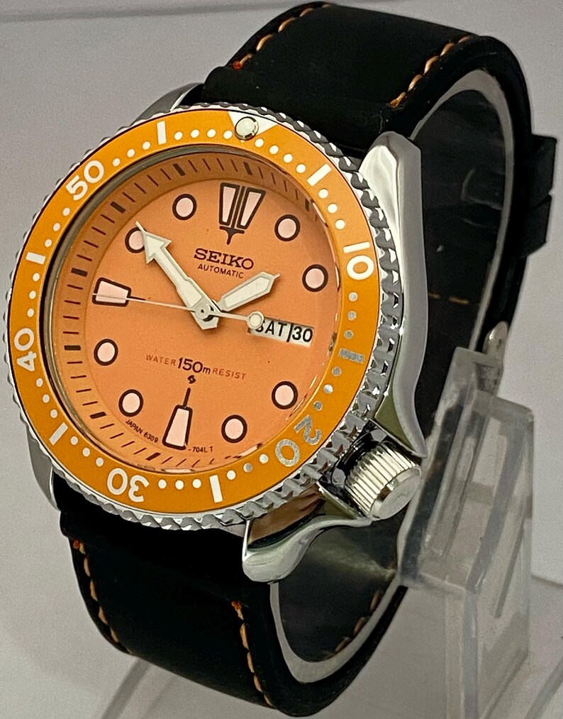 May include: A stainless steel Seiko automatic watch with an orange dial and bezel. The watch features a black leather strap with orange stitching. The dial displays the day and date. The watch is water resistant to 150m.