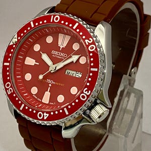 K&ouml;nnte beinhalten: Rote Seiko Automatik-Uhr mit braunem Kautschukarmband. Die Uhr hat ein rotes Zifferblatt mit wei&szlig;en Markierungen und Zeigern, eine drehbare L&uuml;nette und eine Datumsanzeige. Wasserdicht bis 150m.