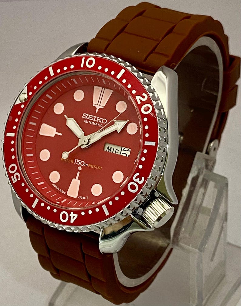 K&ouml;nnte beinhalten: Rote Seiko Automatik-Uhr mit braunem Kautschukarmband. Die Uhr hat ein rotes Zifferblatt mit wei&szlig;en Markierungen und Zeigern, eine drehbare L&uuml;nette und eine Datumsanzeige. Wasserdicht bis 150m.