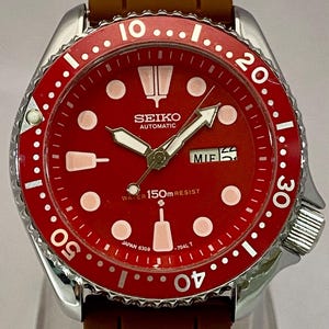 K&ouml;nnte beinhalten: Seiko Automatik-Armbanduhr mit rotem Zifferblatt und braunem Kautschukarmband. Die Uhr hat wei&szlig;e Stundenmarkierungen, Zeiger und eine Datumsanzeige. Die L&uuml;nette ist rot mit wei&szlig;en Markierungen. Wasserdicht bis 150m.