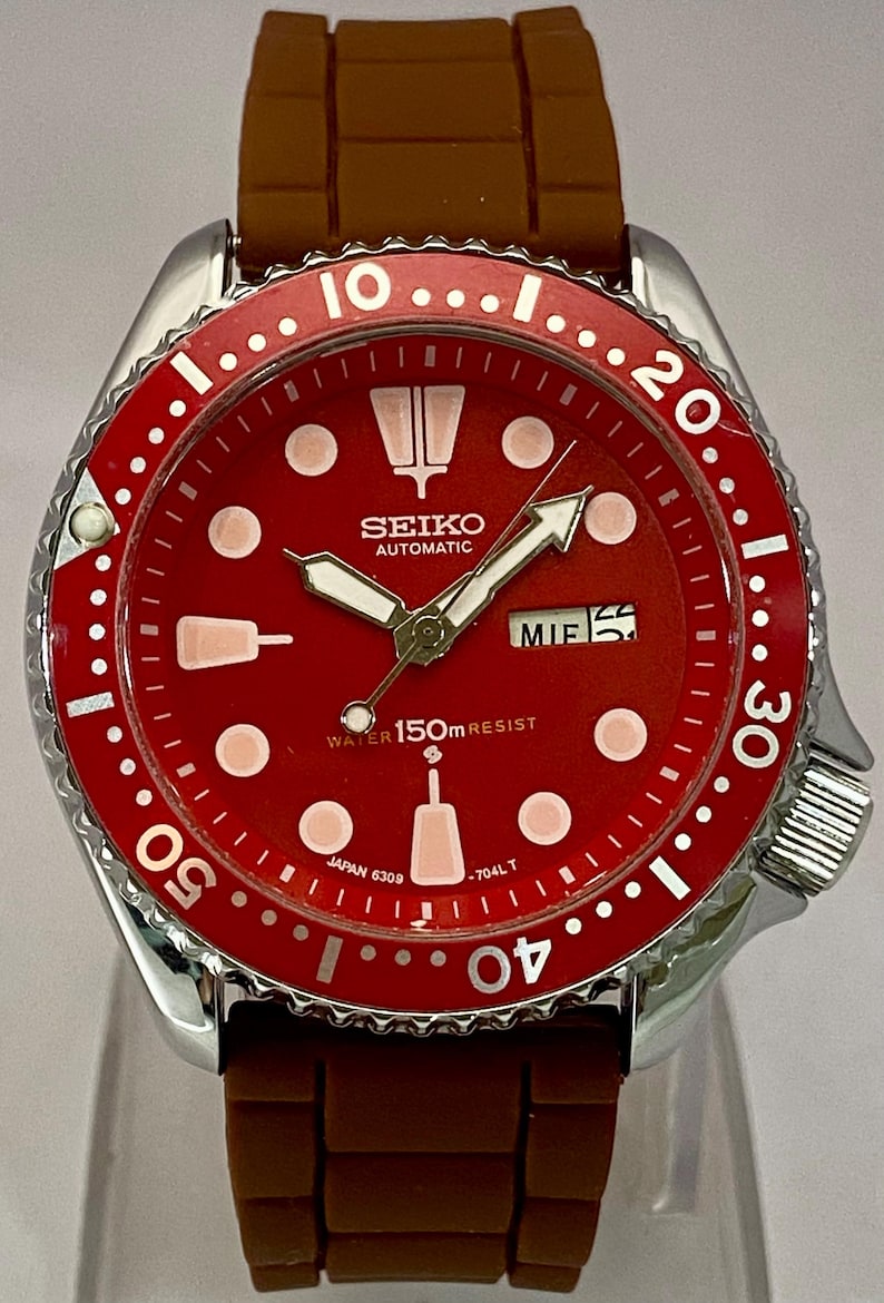 K&ouml;nnte beinhalten: Seiko Automatik-Armbanduhr mit rotem Zifferblatt und braunem Kautschukarmband. Die Uhr hat wei&szlig;e Stundenmarkierungen, Zeiger und eine Datumsanzeige. Die L&uuml;nette ist rot mit wei&szlig;en Markierungen. Wasserdicht bis 150m.