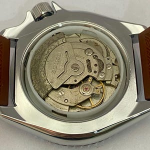 K&ouml;nnte beinhalten: Nahaufnahme eines silberfarbenen Uhrengeh&auml;uses mit braunem Armband. Der transparente Boden zeigt das komplizierte Innenleben, einschlie&szlig;lich Zahnr&auml;dern und der Marke "SEIKO". Die Krone der Uhr ist oben sichtbar.