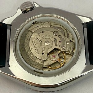 May include: Close-up of a silver-toned watch case with a clear back, revealing the intricate internal mechanism. The watch movement features gears, jewels, and the brand name "SEIKO" prominently displayed. A black leather strap is attached to the watch case.
