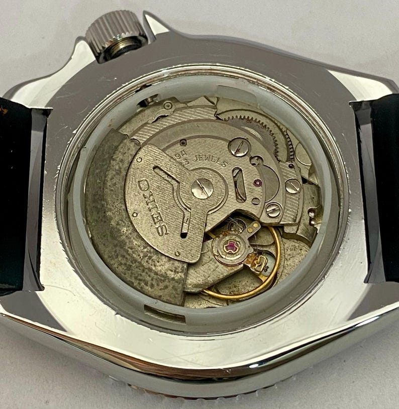 May include: Close-up of a silver-toned watch case with a clear back, revealing the intricate internal mechanism. The watch movement features gears, jewels, and the brand name "SEIKO" prominently displayed. A black leather strap is attached to the watch case.