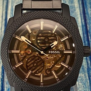 May include: Black Fossil watch with a skeleton dial, showcasing the inner workings of the mechanism. The watch has a black metal band and a textured bezel. The hands and hour markers are gold-toned, and the watch face displays the Fossil logo.