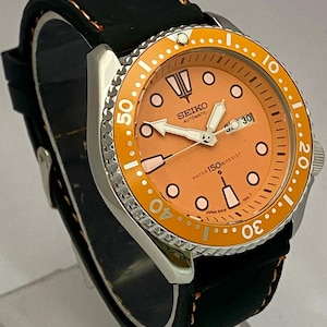 May include: A stainless steel Seiko automatic watch with an orange dial and bezel. The watch features a black leather strap with orange stitching. The dial displays the date and luminous hands and markers. The watch is water resistant to 150 meters.