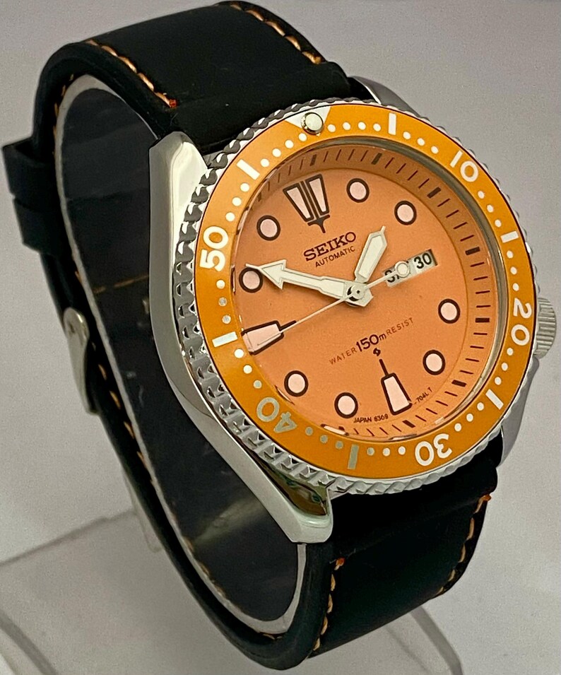 May include: A stainless steel Seiko automatic watch with an orange dial and bezel. The watch features a black leather strap with orange stitching. The dial displays the date and luminous hands and markers. The watch is water resistant to 150 meters.