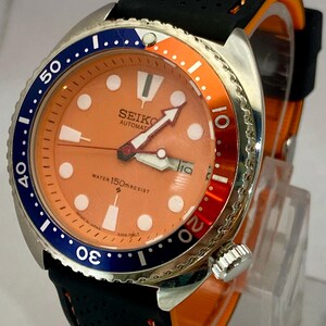 K&ouml;nnte beinhalten: Eine Seiko Automatik-Uhr aus Edelstahl mit orangefarbenem Zifferblatt, blau-oranger L&uuml;nette und schwarzem Armband. Das Zifferblatt zeigt das Datum und den Text "SEIKO AUTOMATIC WATER 150m RESIST".