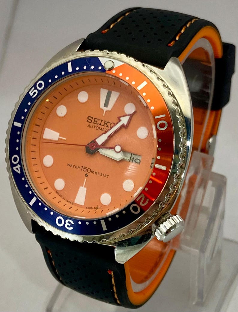 K&ouml;nnte beinhalten: Eine Seiko Automatik-Uhr aus Edelstahl mit orangefarbenem Zifferblatt, blau-oranger L&uuml;nette und schwarzem Armband. Das Zifferblatt zeigt das Datum und den Text "SEIKO AUTOMATIC WATER 150m RESIST".