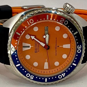 K&ouml;nnte beinhalten: Eine Seiko Automatik-Armbanduhr mit orangefarbenem Zifferblatt und schwarzem Armband. Die Uhr verf&uuml;gt &uuml;ber ein Edelstahlgeh&auml;use, eine drehbare L&uuml;nette mit blauen und roten Akzenten und wei&szlig;e Stundenmarkierungen. Das Armband ist schwarz mit orangefarbenem Futter.