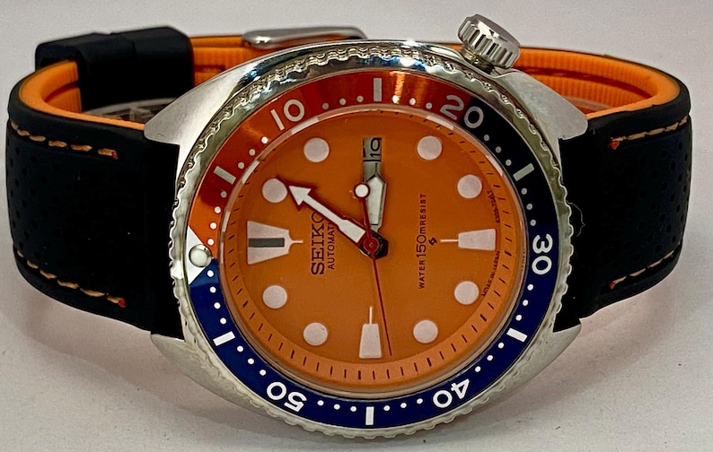 K&ouml;nnte beinhalten: Eine Seiko Automatik-Armbanduhr mit orangefarbenem Zifferblatt und schwarzem Armband. Die Uhr verf&uuml;gt &uuml;ber ein Edelstahlgeh&auml;use, eine drehbare L&uuml;nette mit blauen und roten Akzenten und wei&szlig;e Stundenmarkierungen. Das Armband ist schwarz mit orangefarbenem Futter.