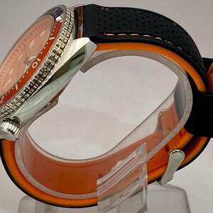 K&ouml;nnte beinhalten: Eine Edelstahl-Armbanduhr mit orangefarbenem Zifferblatt und einem schwarzen und orangefarbenen Armband. Die Uhr hat eine drehbare L&uuml;nette mit blauen und silbernen Markierungen. Das Armband besteht aus schwarzem, perforiertem Gummi und orangefarbenem Stoff.