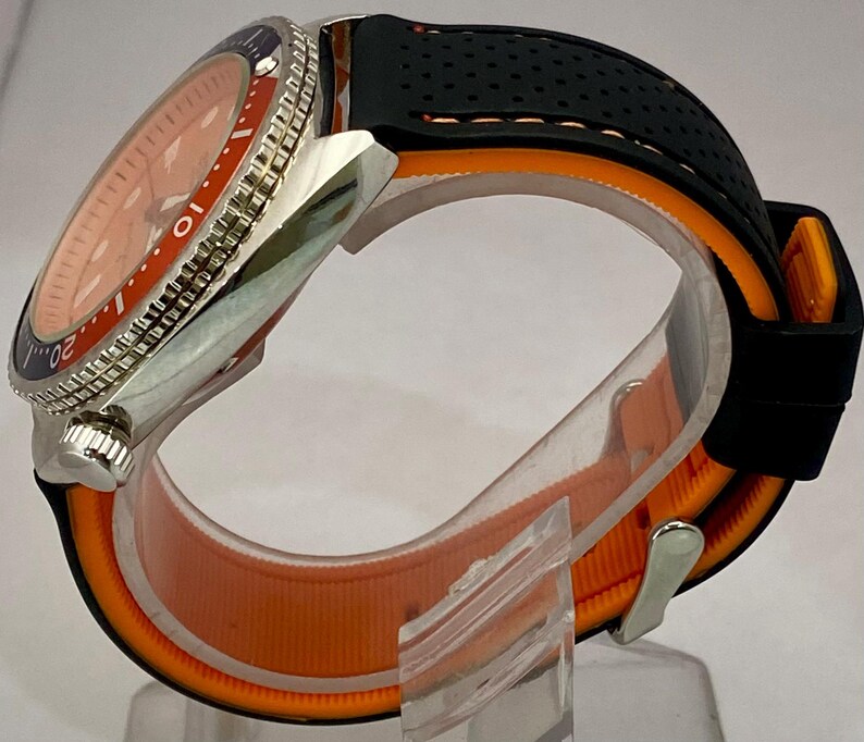 K&ouml;nnte beinhalten: Eine Edelstahl-Armbanduhr mit orangefarbenem Zifferblatt und einem schwarzen und orangefarbenen Armband. Die Uhr hat eine drehbare L&uuml;nette mit blauen und silbernen Markierungen. Das Armband besteht aus schwarzem, perforiertem Gummi und orangefarbenem Stoff.