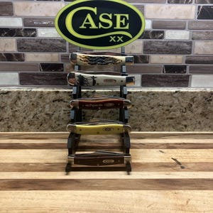 May include: A black display stand holding five Case pocket knives. The knives have various handle designs and colors. A sign with the word "CASE" in yellow and white is at the top of the stand.