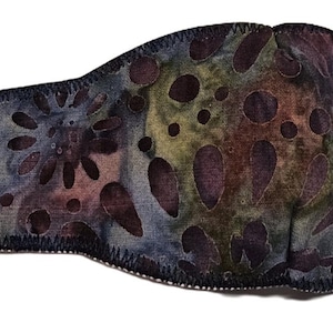 May include: A fabric eye patch with a batik-style pattern in shades of blue, purple, and green. The patch features a floral and dot design, a black elastic strap, and a black border. The eye patch is designed to cover one eye.