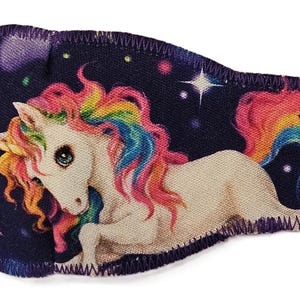 May include: A whimsical eye patch featuring a white unicorn with a rainbow mane and tail, set against a dark blue starry background. The patch has a purple border and a pink elastic strap.