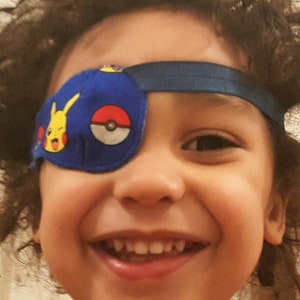 May include: A young child with curly brown hair is wearing a blue eye patch with a cartoon character on it. The child is smiling and showing their teeth.