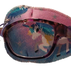 Pode incluir: Óculos roxos e tartaruga com um tapa-olho com estampa de unicórnio. O tapa-olho é rosa e azul com um unicórnio e estrelas.